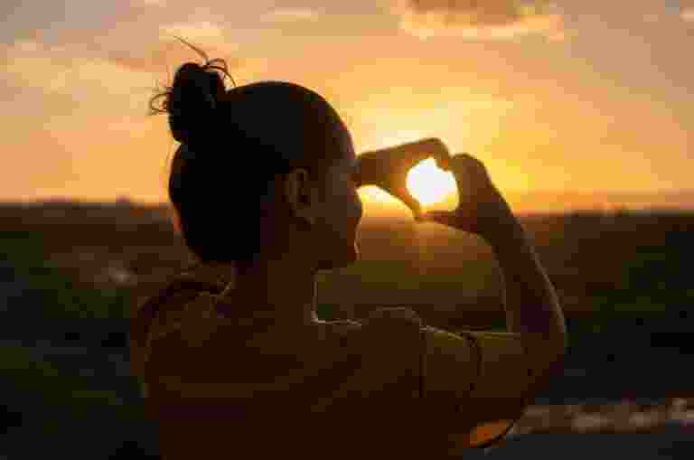 A person makes a love heart shape from their hands and holds it up to an orange and yellow sunset.