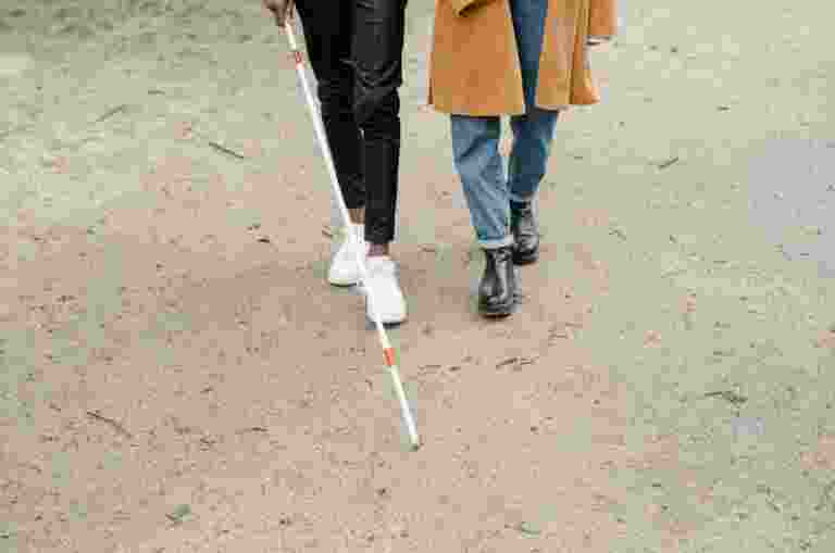 Two people walk along the beach together. One person is holding a White-Cane