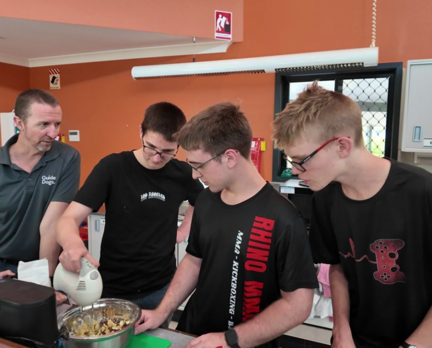 Three teens and a Guide Dogs instructor prepare food together, one using a hand mixer over a bowl.