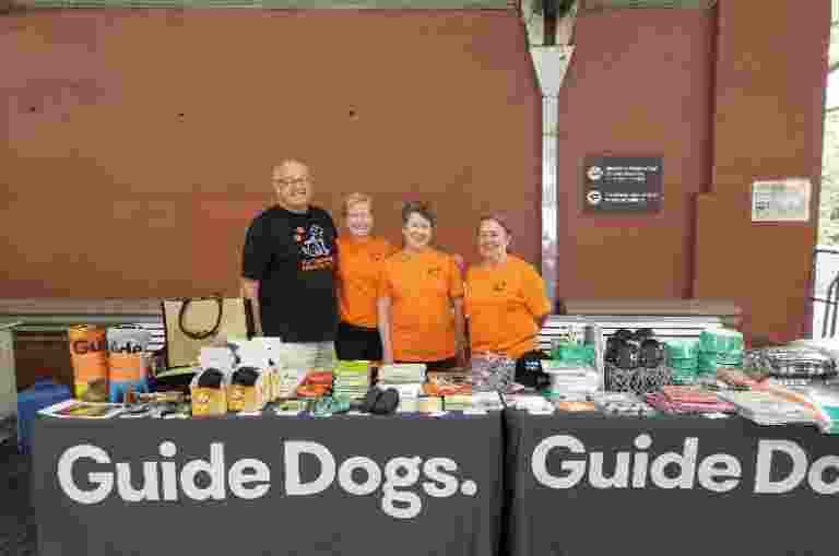 Four people standing behind a Guide Dogs merchandise table wearing Guide Dogs shirts.