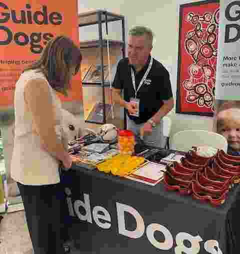 Two people standing at a Guide Dogs stall. The woman is looking at products on the table.