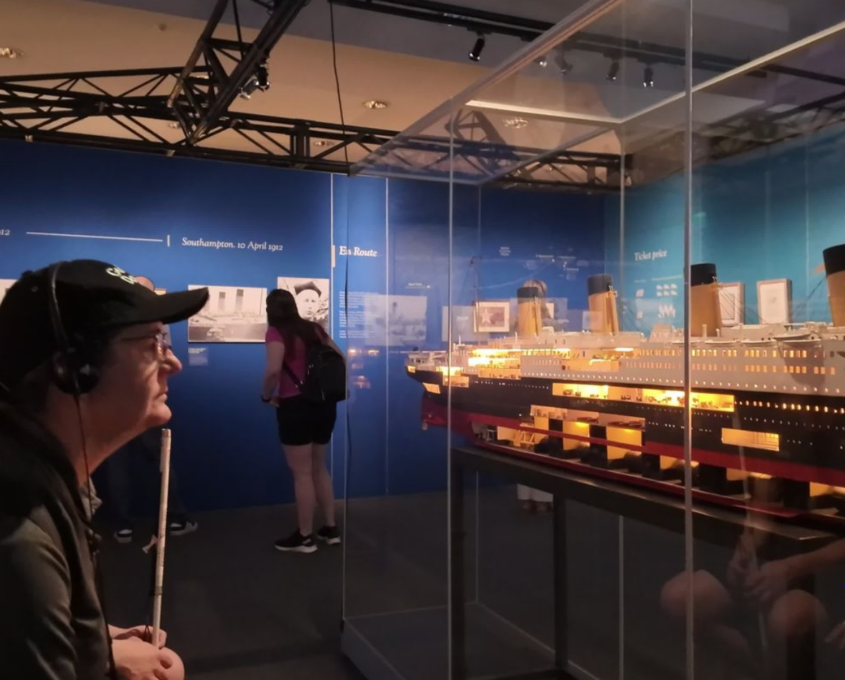 Man with a white cane and headset observing a detailed, illuminated model of the Titanic in a museum exhibit.