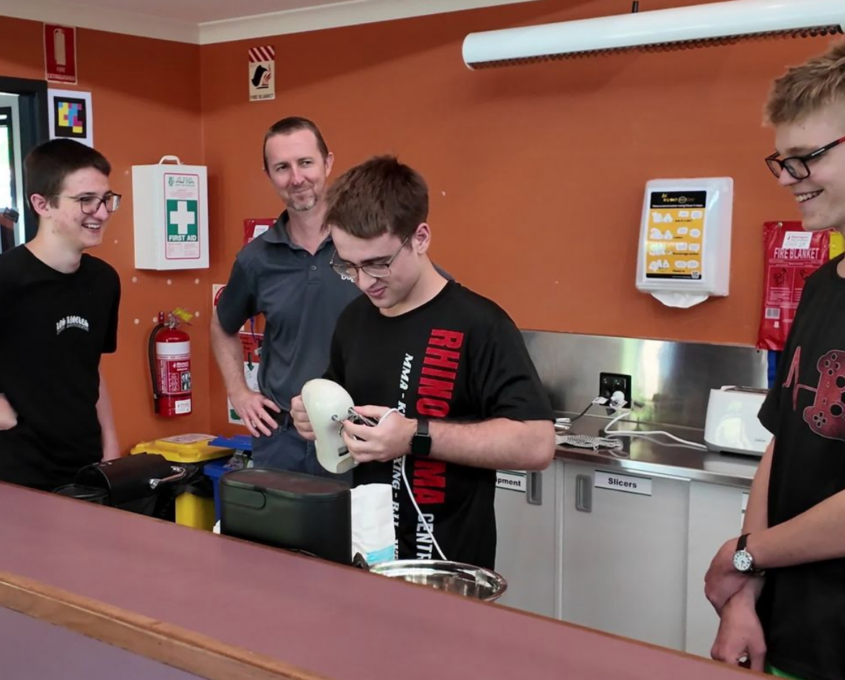 Teen boy examines an electric hand mixer while others and an instructor smile nearby in a kitchen classroom.
