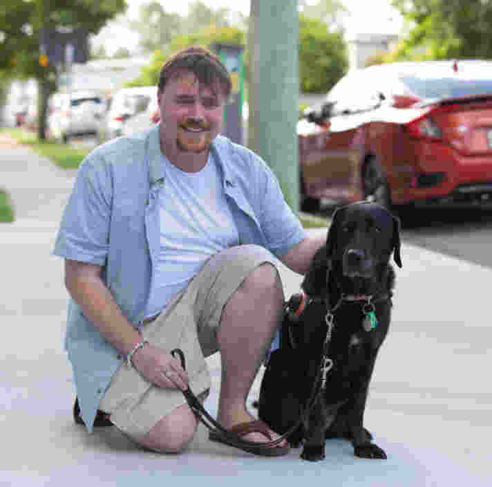 Image shows Guide Dog Handler Chris and Guide Dog Alyssa sit on a footpath together, smiling to the camera.