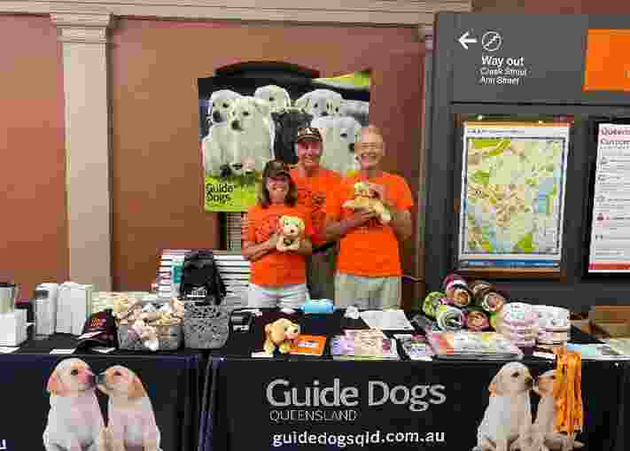 Image shows Guide Dogs volunteers standing at a merchandise stall, smiling at the camera.
