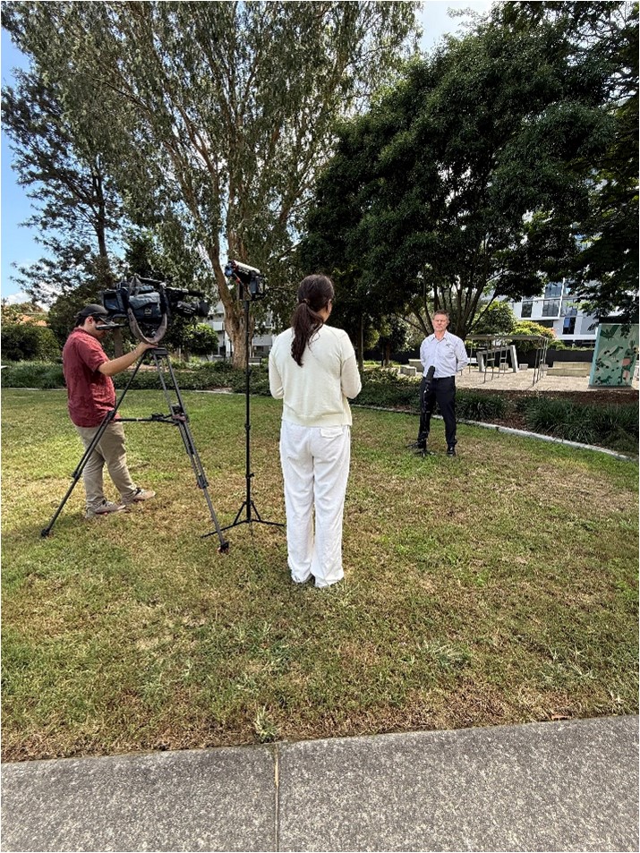 Image shows a news crew filming an outdoor interview in a park, with a woman facing a man speaking on camera as a videographer records the scene.