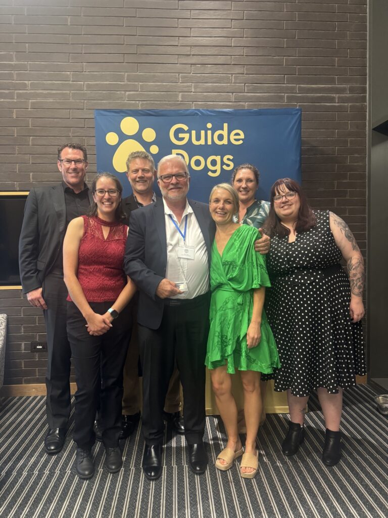 Image shows Marc Lyell and a group of smiling Guide Dogs team members standing together at the IGDF conference, commemorating Marc’s receipt of the prestigious Ken Lord Award for decades of innovation and leadership in mobility training.