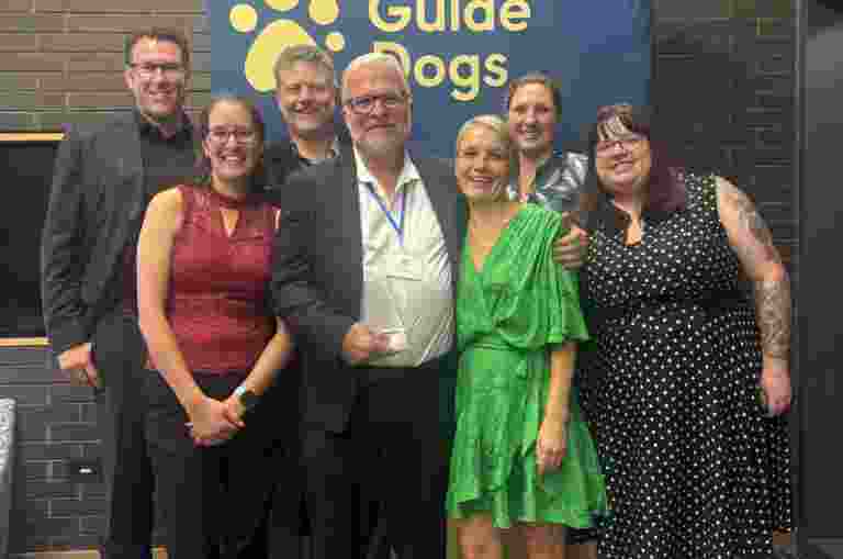 Image shows Marc Lyell and a group of smiling Guide Dogs team members standing together at the IGDF conference, commemorating Marc’s receipt of the prestigious Ken Lord Award for decades of innovation and leadership in mobility training.