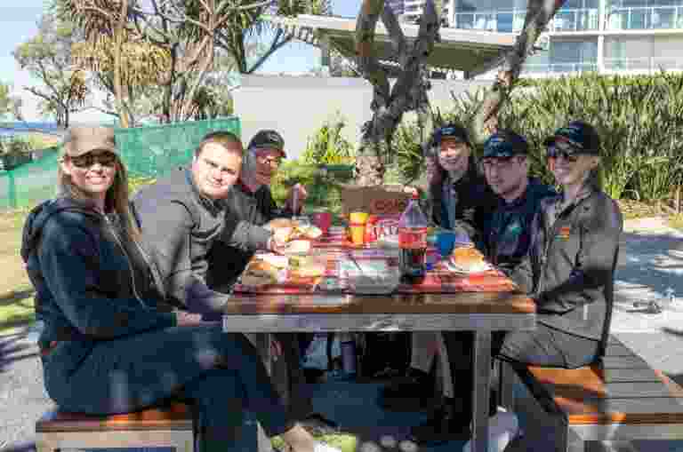 Image shows six smiling campers and staff sitting at an outdoor picnic table, enjoying lunch together on the final day of the Guide Dogs Mobility Camp. A red-checked tablecloth holds soft drinks, sandwiches, and snacks.