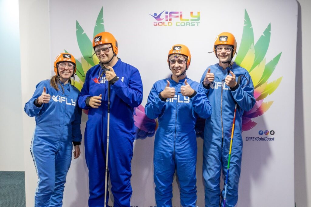Image shows four young participants at iFly Gold Coast, smiling in blue flight suits and orange helmets during a Guide Dogs Queensland Mobility Camp. They stand in front of a colorful wing backdrop, giving thumbs-up after indoor skydiving.