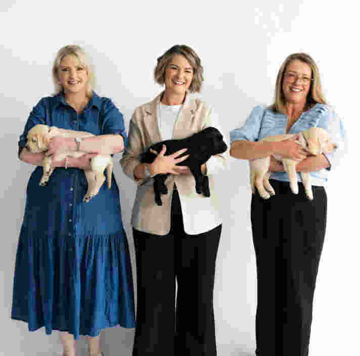Image shows Corinne (Left), Renae (Middle) and Liz (Right) holding Labrador puppies and smiling to the camera.