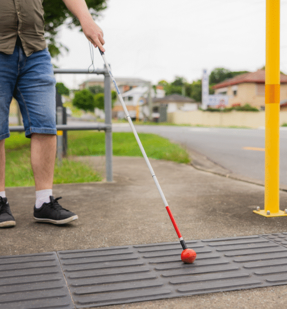 Image shows a person using a white cane with a red tip to navigate tactile paving at a street crossing, indicating an accessible pedestrian path.