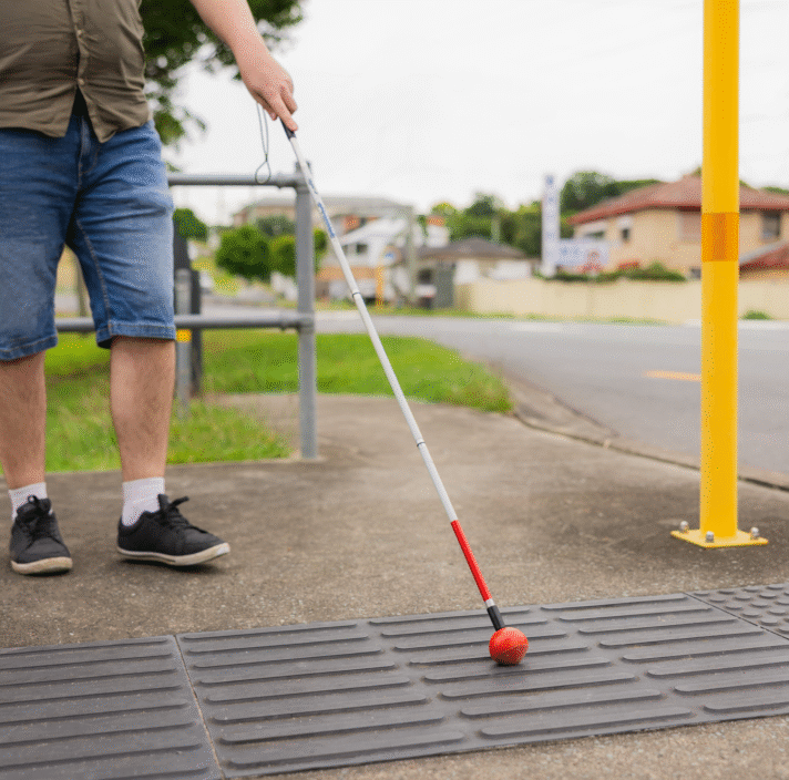Image shows a person using a white cane with a red tip to navigate tactile paving at a street crossing, indicating an accessible pedestrian path.