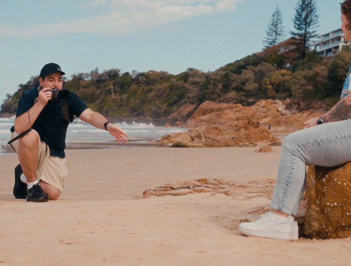 Image shows James, a photographer with low vision, capturing a portrait on the beach; he’s using a white cane and gesturing toward the subject sitting on a large rock.