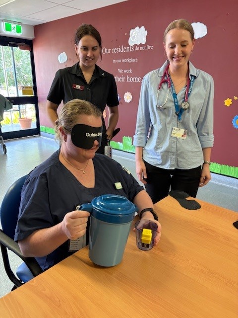 A staff member wearing a blindfold practices handling a jug while two colleagues observe.
