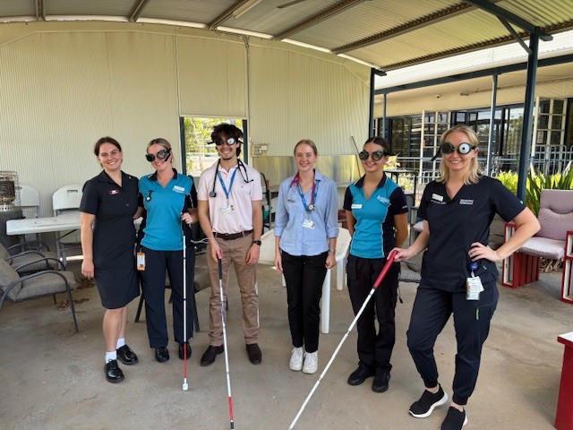 Six hospital staff pose outdoors, with two wearing low-vision goggles and holding white canes.