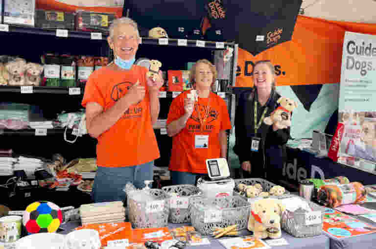 Three smiling volunteers stand behind a crowded merchandise stall for Guide Dogs Queensland at an outdoor event. Two volunteers on the left wear bright orange "Paws Up for Guide Dogs" T-shirts, with the man on the far left giving a thumbs up and holding a small plush puppy.
