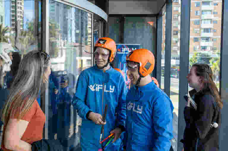 Two young men in blue iFLY jumpsuits and orange helmets stand together, smiling and talking. They are both holding white long canes. A woman in an orange shirt and another woman in a black jacket stand on either side of them, also engaged in the conversation. They are standing in an indoor skydiving facility next to the glass flight chamber.