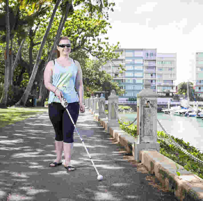 A female in walking attire is using a white cane while walking along a path next to a river. There are buildings in the background and boats in the water.