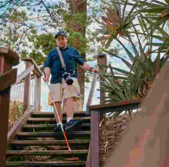 A man wearing a camera and a cap uses a long white cane to navigate down a set of outdoor wooden stairs. The scene is surrounded by coastal greenery and trees under a blue sky.