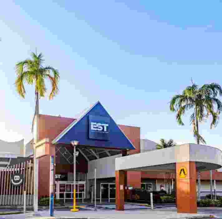 An exterior view of Earlville Shopping Town entrance. The layout is spacious and flat, showing a clear path from the bus stop and pedestrian crossing to the automatic glass doors under a large blue triangular awning.