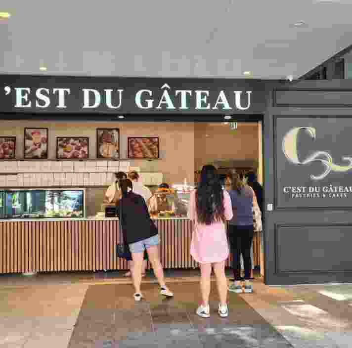 A street-level view of C'est Du Gateau bakery. The shop has a wide, step-free entrance and a long service counter. Several customers stand on the spacious, even-tiled pavement, highlighting the accessible and open floor plan.