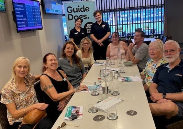 A group of participants and staff sit around a long table for a Guide Dogs Queensland workshop in a community venue with digital screens and banners.