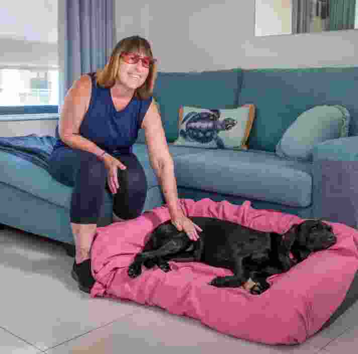 Image shows Penny, a Guide Dogs Client sitting in a blue chair. Next to her is her black Guide Dog Kiki, laying on a pink bed.