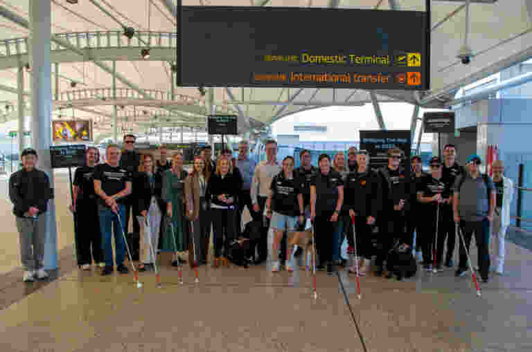 A large group of people, including individuals holding white canes and some accompanied by guide dogs, stand together posing for a group photo at an open-air airport terminal. Above them, a large digital sign points toward the "Domestic Terminal" and "International transfer." Several people hold small signs that read "Bridging The Gap to 2032."