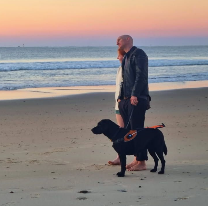 Guide dogs client; Rhett standing on the beach during a sunset, with his black Guide Dog next to him.