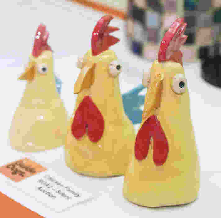 Three small, handmade ceramic chicken figurines standing in a row on a white surface. The yellow, cone-shaped chickens feature bright red combs and wattles, wide, cartoonish eyes, and small orange beaks. A small printed sign next to them reads "Guide Dogs" and "Chicken Family."