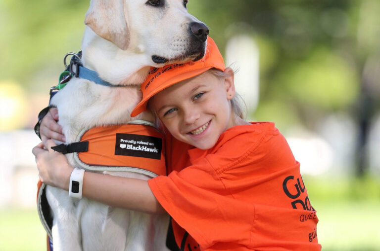 A smiling young girl wearing an orange t-shirt and cap sits on the grass, warmly hugging a white Labrador guide dog wearing an orange vest.
