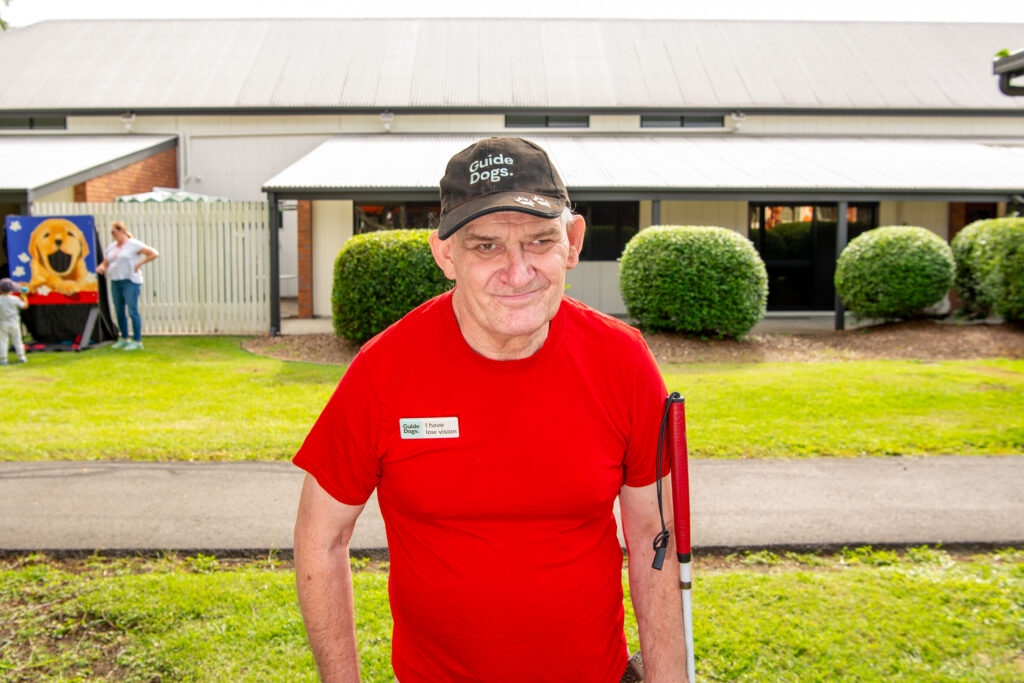 A smiling man named Ian wears a red shirt and a Guide Dogs cap while holding his white cane on the Guide Dogs Queensland campus.