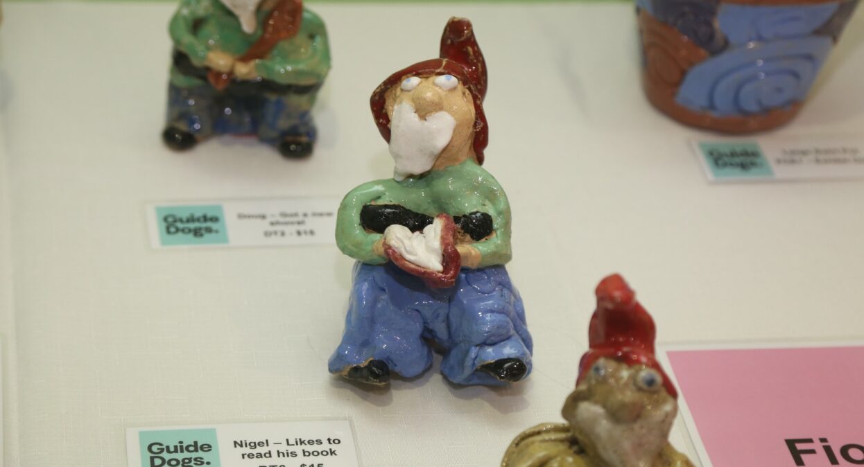 A close-up shot of small, colorful handmade ceramic garden gnome figurines displayed with "Guide Dogs" labels on a white tablecloth.