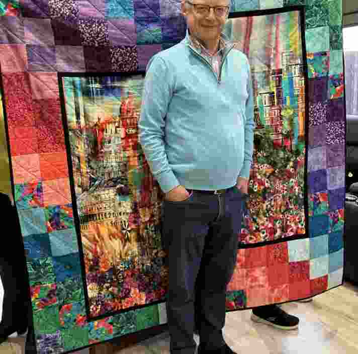 A man standing in front of a patchwork quilt, smiling at the camera.