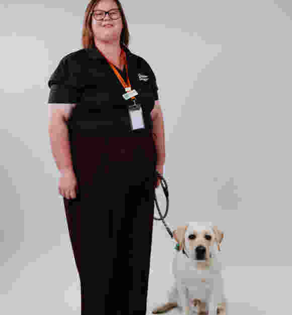 Gemma standing in her black Guide Dog uniform, smiling, as Keith, yellow ambassador Labrador, sits at her feet.